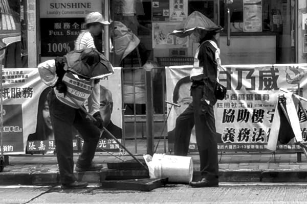 Deux agents de propreté au travail dans une rue de Hong Kong, devant une palissade couverte d'affiches en chinois.